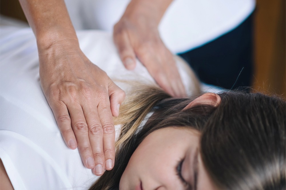 Reiki: Behandlung mit zwei Händen im Schulterbereich bei Frau in Bauchlage